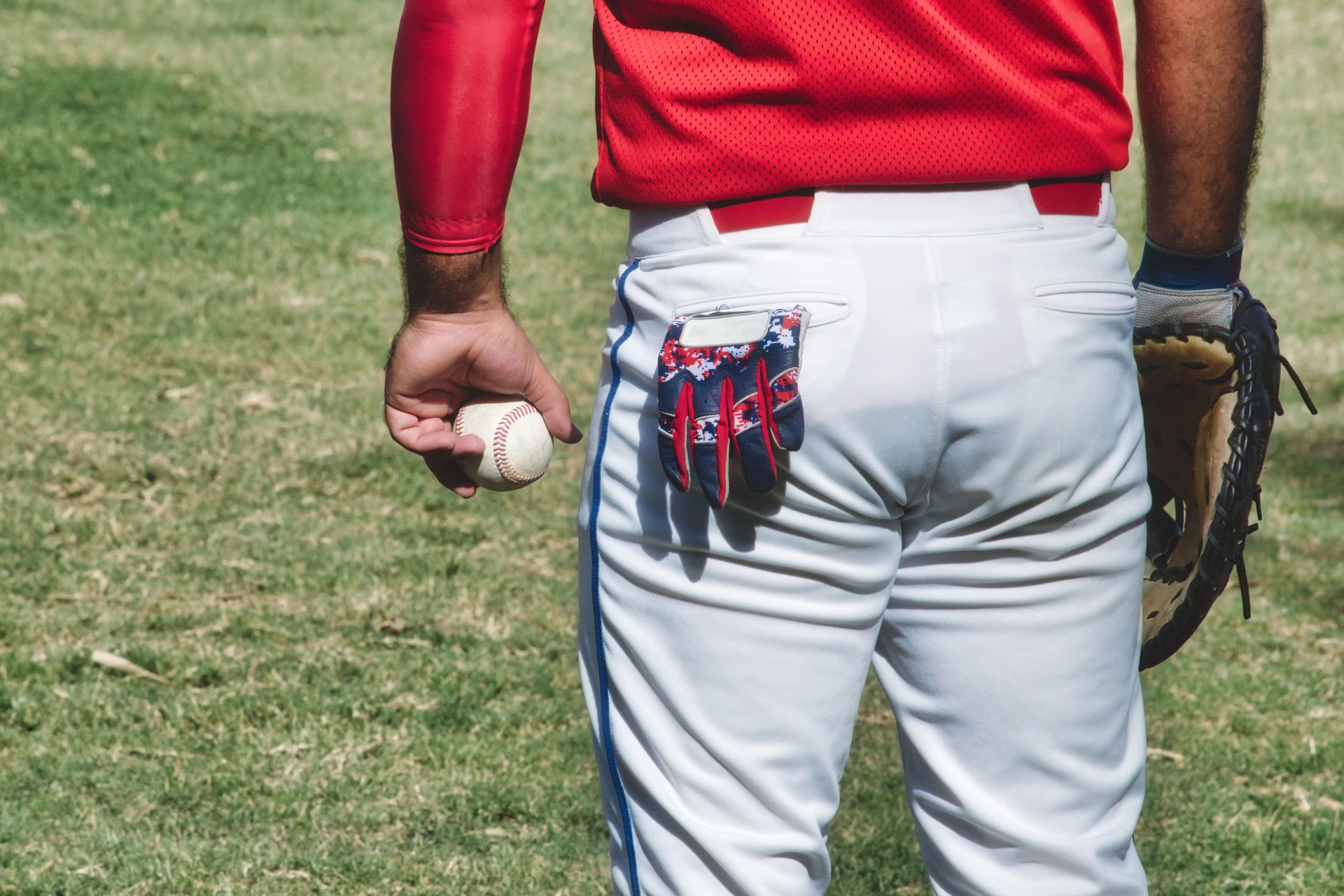 A baseball player at a match wearing a leather mitt and holding a ball in his hand, with a small glove sticking out of his back pocket in white pants, standing on the grass